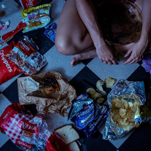 Man sitting surrounded by assorted snacks and packaging on a checkered floor, depicting a binge-eating scene.