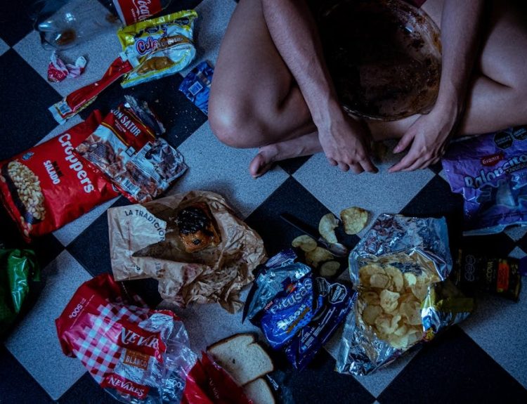 Man sitting surrounded by assorted snacks and packaging on a checkered floor, depicting a binge-eating scene.